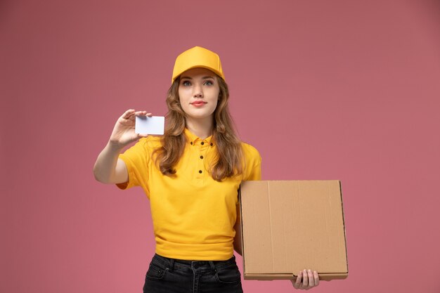 Giovane corriere femminile di vista frontale in uniforme gialla che tiene carta bianca e pacchetto di cibo sul lavoratore di servizio di lavoro di consegna uniforme scrivania rosa scuro