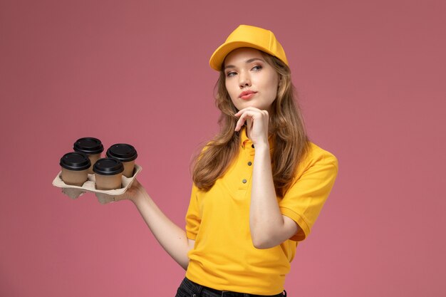 Giovane corriere femminile di vista frontale in uniforme gialla che tiene i bicchieri di plastica del caffè marrone che pensa sulla lavoratrice di servizio di consegna dell'uniforme della scrivania rosa scuro