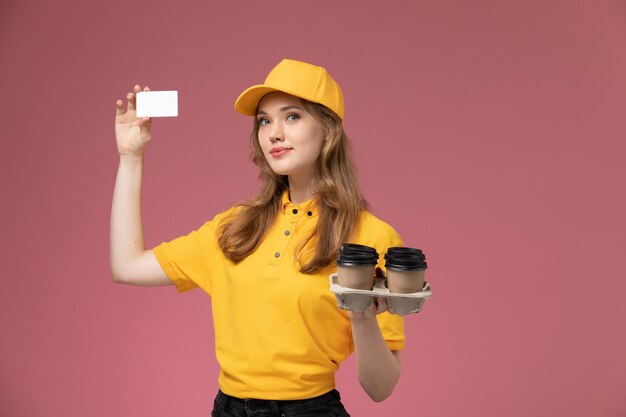 Giovane corriere femminile di vista frontale in uniforme gialla che tiene i bicchieri di plastica del caffè marrone con la carta sul lavoratore femminile di servizio di consegna dell'uniforme dello scrittorio rosa scuro