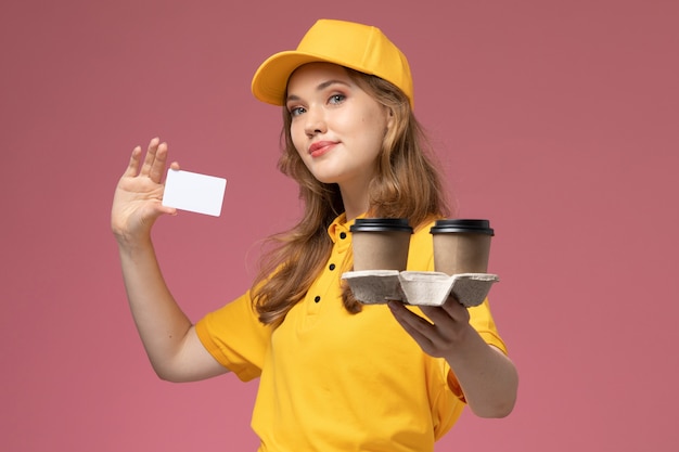 Giovane corriere femminile di vista frontale in uniforme gialla che tiene le tazze di caffè di plastica e carta bianca sul lavoratore di servizio di consegna dell'uniforme dello scrittorio rosa scuro