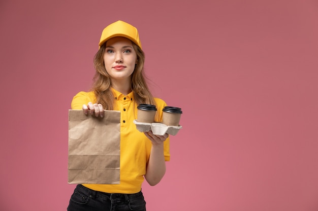 Giovane corriere femminile di vista frontale in uniforme gialla che tiene le tazze di caffè e il pacchetto di cibo sul lavoratore di servizio di lavoro di consegna uniforme da scrivania rosa scuro