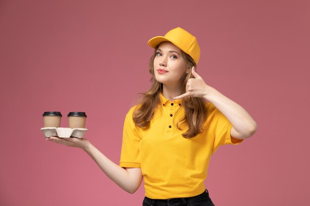Giovane corriere femminile di vista frontale in uniforme gialla che tiene le tazze di caffè marroni di plastica che posano sul lavoratore di servizio di colore di consegna uniforme di lavoro del fondo rosa