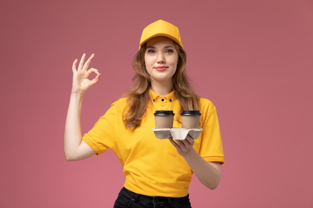 Giovane corriere femminile di vista frontale in uniforme gialla che tiene le tazze di caffè sul lavoratore rosa scuro del servizio di consegna dell'uniforme di lavoro della scrivania dello scrittorio