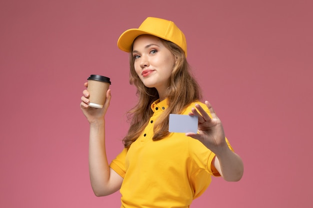 Giovane corriere femminile di vista frontale in uniforme gialla che tiene tazza di caffè e carta grigia sul lavoratore di servizio di lavoro di consegna uniforme scrivania rosa scuro