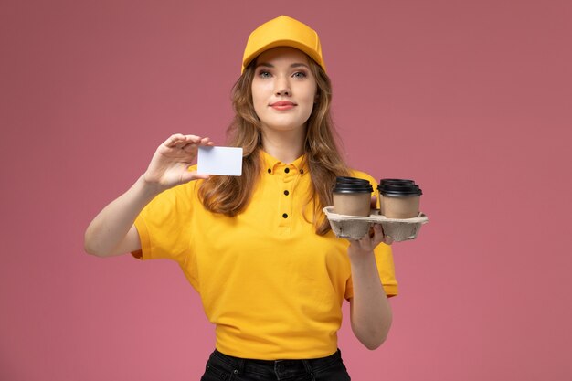Giovane corriere femminile di vista frontale in uniforme gialla che tiene tazze di caffè e carta bianca sul lavoratore di servizio di lavoro di consegna uniforme da scrivania rosa scuro
