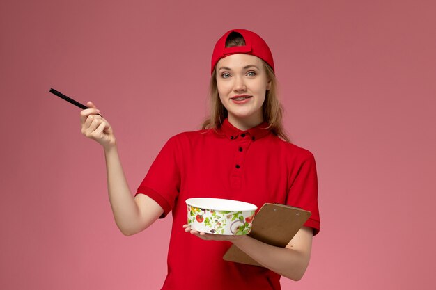 Giovane corriere femminile di vista frontale in uniforme rossa e ciotola di consegna della tenuta del capo e blocco note con la parete rosa della penna