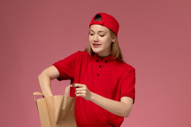 Giovane corriere femminile di vista frontale in uniforme rossa e mantello che tiene il pacchetto di cibo di consegna prendendo qualcosa dall'interno sulla parete rosa