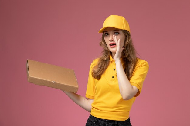 Giovane corriere femminile di vista frontale nel mantello giallo uniforme giallo che tiene la scatola di consegna del cibo sulla lavoratrice di servizio di consegna uniforme sfondo rosa scuro