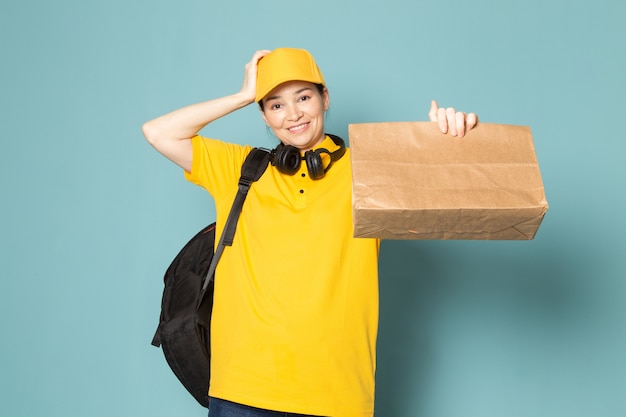 giovane corriere femminile in t-shirt gialla tappo giallo tenendo la scatola sulla parete blu