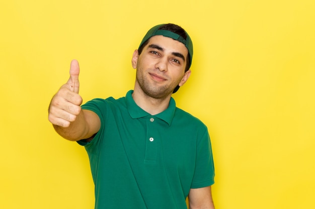 Giovane corriere maschio di vista frontale in berretto verde della camicia verde che sorride e che mostra il segno impressionante sul colore del servizio di consegna del lavoro del fondo giallo