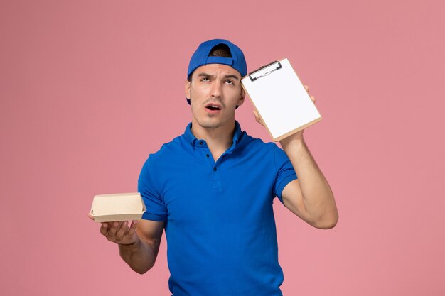 Giovane corriere maschio di vista frontale in capo uniforme blu che pensa e che tiene poco pacchetto di cibo di consegna e blocco note sulla parete rosa chiaro