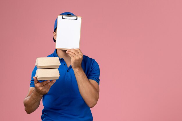 Giovane corriere maschio di vista frontale in capo uniforme blu che tiene i piccoli pacchetti dell'alimento di consegna con il blocco note sulla parete rosa