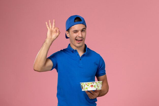 Giovane corriere maschio di vista frontale in capo uniforme blu che tiene la ciotola rotonda di consegna che sbatte le palpebre sulla parete rosa-chiaro