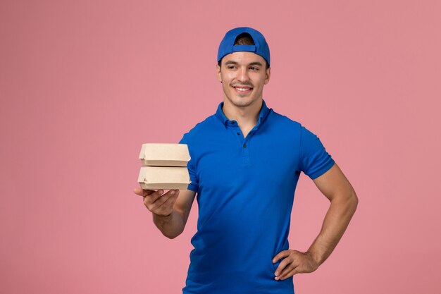 Giovane corriere maschio di vista frontale in capo uniforme blu che tiene piccoli pacchetti dell'alimento di consegna sulla parete rosa