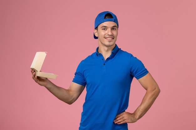 Giovane corriere maschio di vista frontale in capo uniforme blu che tiene piccoli pacchetti dell'alimento di consegna sulla parete rosa