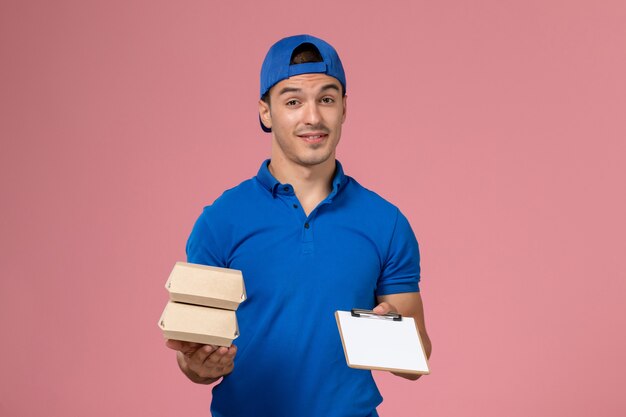 Giovane corriere maschio di vista frontale in capo uniforme blu che tiene piccoli pacchetti di cibo di consegna con il blocco note sulla parete rosa-chiaro