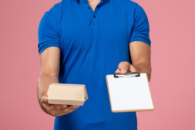 Giovane corriere maschio di vista frontale in capo uniforme blu che tiene poco pacchetto dell'alimento di consegna e blocchetto per appunti sulla parete rosa-chiaro