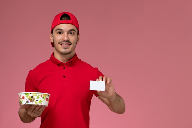 Giovane corriere maschio di vista frontale in capo uniforme rosso che tiene carta di plastica bianca e ciotola di consegna sui precedenti rosa-chiaro.