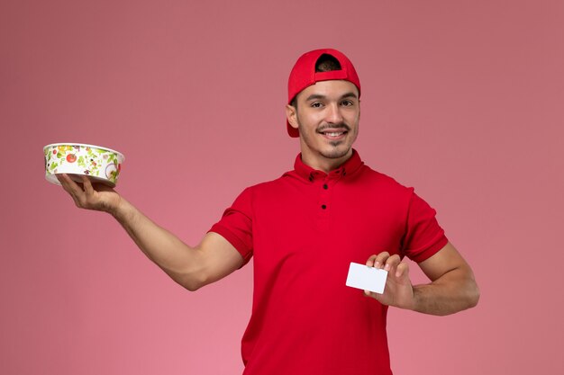 Giovane corriere maschio di vista frontale in capo uniforme rosso che tiene carta di plastica bianca e ciotola di consegna sui precedenti rosa-chiaro.