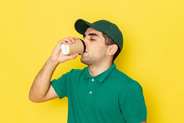 Giovane corriere maschio di vista frontale in cappuccio verde della camicia verde che beve caffè su colore giallo