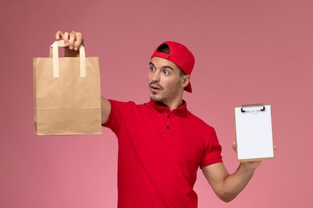 Giovane corriere maschio di vista frontale in mantello rosso uniforme che tiene il pacchetto alimentare e blocco note sorridente sullo sfondo rosa.