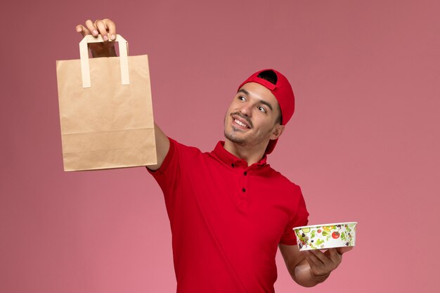 Giovane corriere maschio di vista frontale in mantello rosso uniforme che tiene il pacchetto alimentare e la ciotola sullo sfondo rosa.