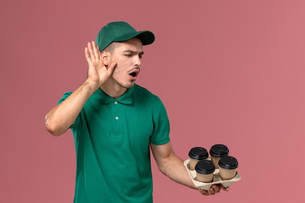 Giovane corriere maschio di vista frontale in uniforme verde che tiene le tazze di caffè marroni che ascolta attentamente su fondo rosa-chiaro