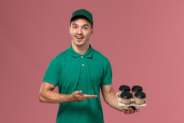 Giovane corriere maschio di vista frontale in uniforme verde che tiene le tazze di caffè marroni che sorridono su fondo rosa-chiaro