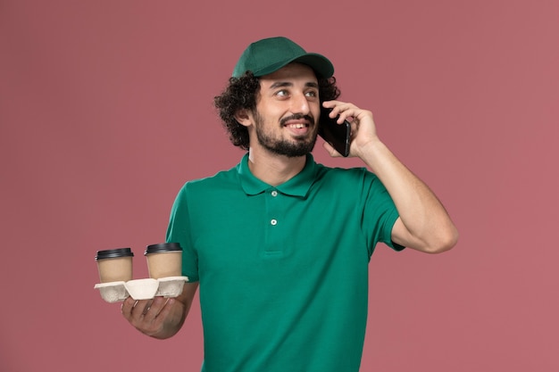 Giovane corriere maschio di vista frontale in uniforme verde e mantello che tiene le tazze di caffè di consegna che parlano sul lavoratore di consegna uniforme di lavoro di sfondo rosa