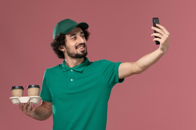 Giovane corriere maschio di vista frontale in uniforme verde e mantello che tiene le tazze di caffè di consegna che prendono la foto sull'operaio di consegna dell'uniforme di lavoro di servizio del fondo rosa