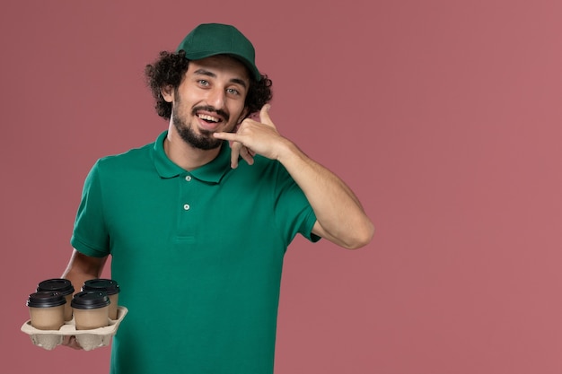 Giovane corriere maschio di vista frontale in uniforme verde e mantello che tiene le tazze di caffè di consegna marrone che sorridono sul lavoro di lavoro di consegna uniforme di servizio del fondo rosa