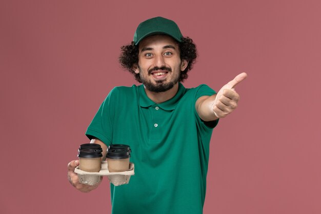 Giovane corriere maschio di vista frontale in uniforme verde e mantello che tiene le tazze di caffè di consegna marroni sul lavoro di consegna uniforme di servizio sfondo rosa chiaro