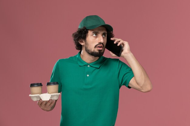 Giovane corriere maschio di vista frontale in uniforme verde e mantello che tiene le tazze di caffè di consegna sull'operaio di consegna dell'uniforme di servizio di sfondo rosa