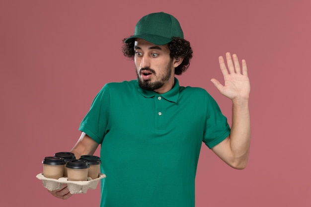 Giovane corriere maschio di vista frontale in uniforme verde e mantello che tiene le tazze di caffè marroni di consegna sull'operaio di consegna dell'uniforme di servizio del fondo rosa chiaro