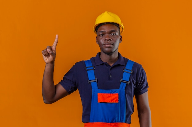 Giovane costruttore afroamericano che indossa l'uniforme da costruzione e casco di sicurezza che mostra e indica con il dito numero uno che sembra fiducioso sul rosa isolato