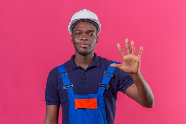 Giovane costruttore afroamericano che indossa l'uniforme da costruzione e casco di sicurezza che mostra e rivolto verso l'alto con le dita numero cinque mentre sorride fiducioso sul rosa isolato