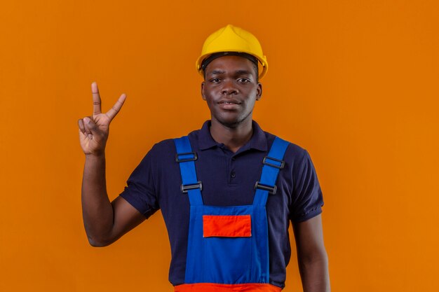 Giovane costruttore afroamericano che indossa l'uniforme da costruzione e casco di sicurezza che mostra e rivolto verso l'alto con le dita numero due mentre sorride fiducioso sull'arancio isolato
