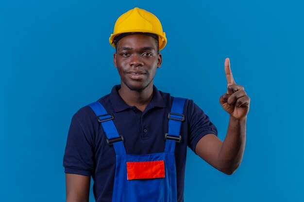 Giovane costruttore afroamericano che indossa l'uniforme da costruzione e casco di sicurezza che punta il dito verso l'alto con espressione fiduciosa in piedi sul blu