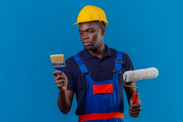 Giovane costruttore afroamericano che indossa l'uniforme da costruzione e casco di sicurezza che tiene pennello e rullo di vernice guardando il pennello con espressione seria in piedi sul blu