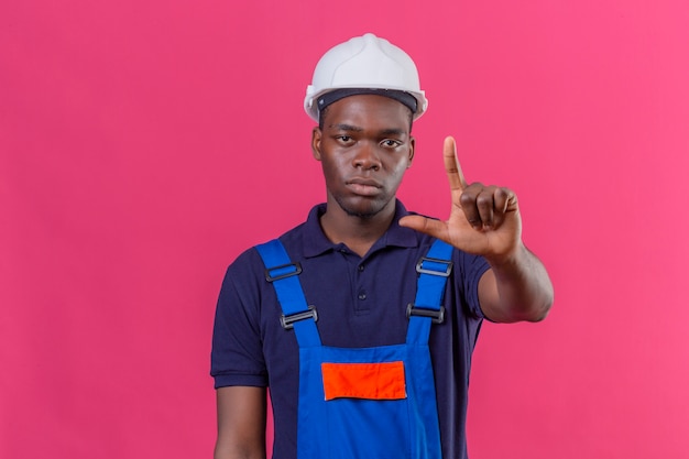 Giovane costruttore afroamericano che indossa l'uniforme da costruzione e casco di sicurezza in piedi con il dito in alto avvertimento di pericolo sul rosa isolato