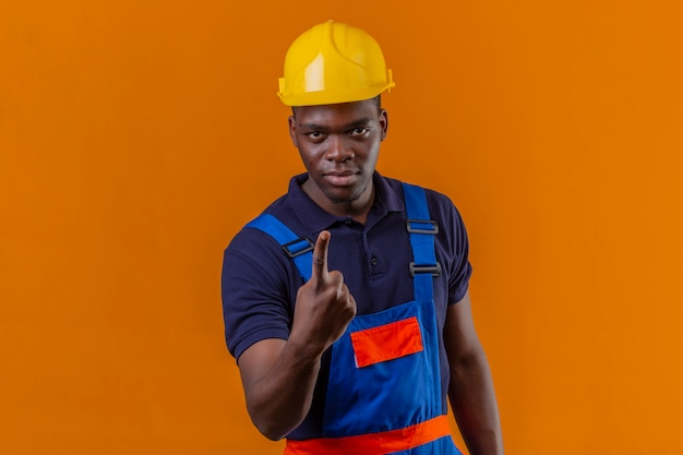 Giovane costruttore afroamericano che indossa l'uniforme da costruzione e casco di sicurezza rivolto verso l'alto con il dito indice che mostra il numero uno guardando con espressione seria fiduciosa in piedi