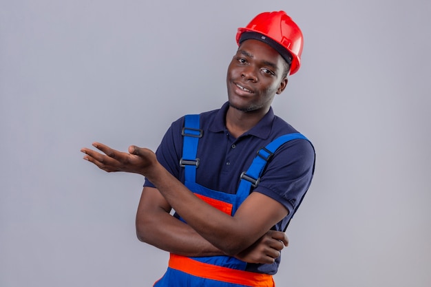 Giovane costruttore afroamericano che indossa l'uniforme da costruzione e il casco di sicurezza che punta con il palmo della mano con il sorriso in piedi