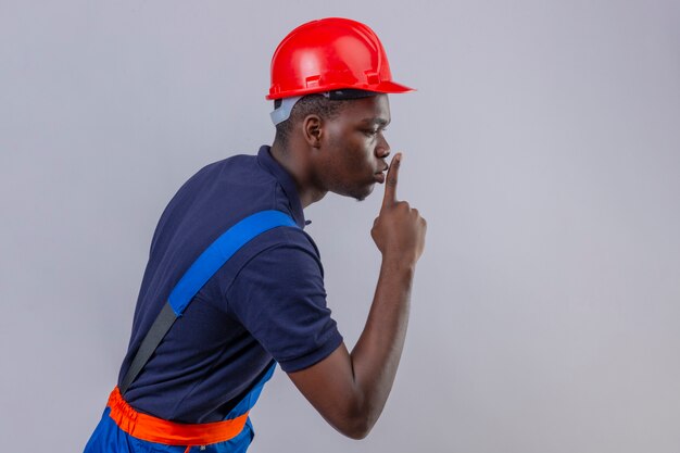Giovane costruttore afroamericano che indossa l'uniforme da costruzione e il casco di sicurezza in piedi lateralmente chiedendo di tacere con il dito sulle labbra silenzio e concetto segreto