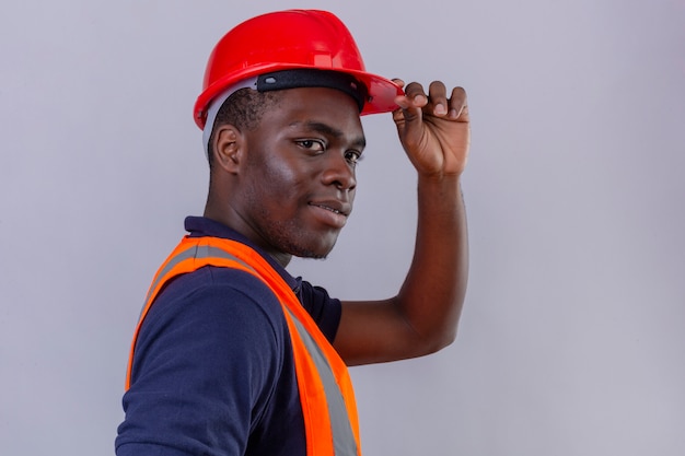 Giovane costruttore afroamericano che indossa la maglia della costruzione e il casco di sicurezza con il casco commovente sicuro di sorriso