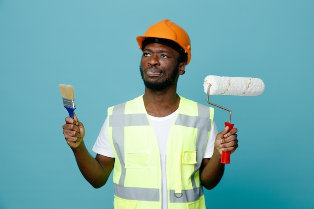 giovane costruttore afroamericano di pensiero in uniforme che tiene una spazzola a rullo con pennello isolato su sfondo blu