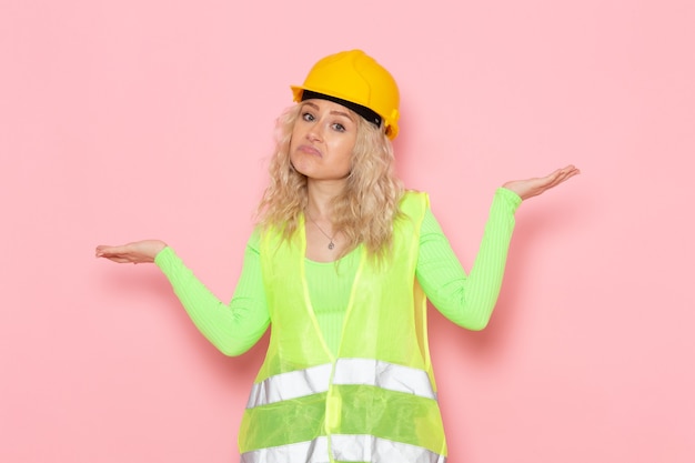 Giovane costruttore femminile di vista frontale in casco giallo del vestito verde della costruzione che posa con l'espressione confusa sulla foto rosa della costruzione di architettura di lavoro dello spazio