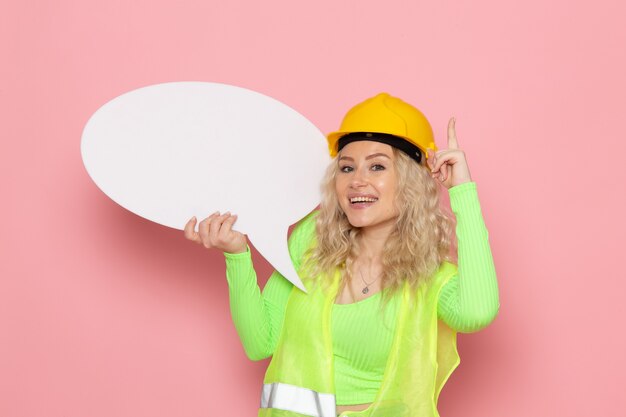 Giovane costruttore femminile di vista frontale nel casco giallo del vestito verde della costruzione che tiene grande segno bianco sulla foto della costruzione di architettura di lavoro dello spazio rosa