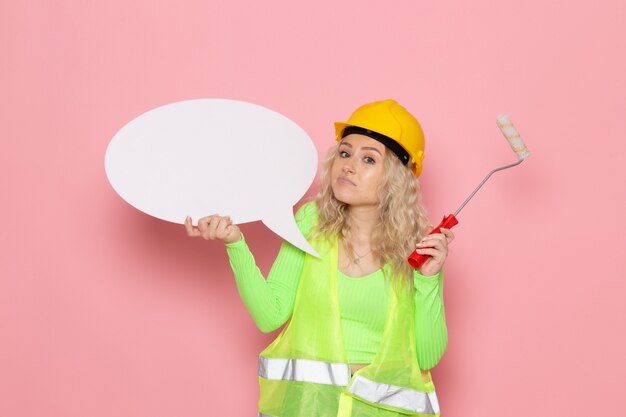 Giovane costruttore femminile di vista frontale nel casco verde del vestito della costruzione che tiene segno e spazzola bianchi sullo spazio rosa