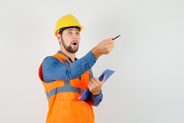 Giovane costruttore in camicia, gilet, casco che dà istruzioni mentre si tiene appunti, vista frontale.