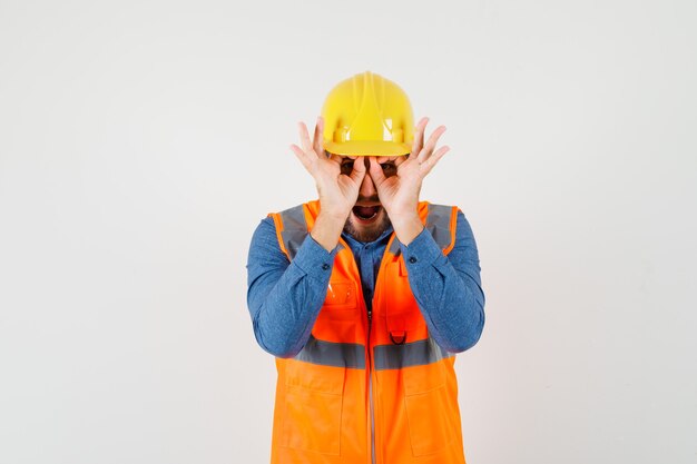 Giovane costruttore in camicia, gilet, casco che mostra il gesto degli occhiali e sembra divertente, vista frontale.